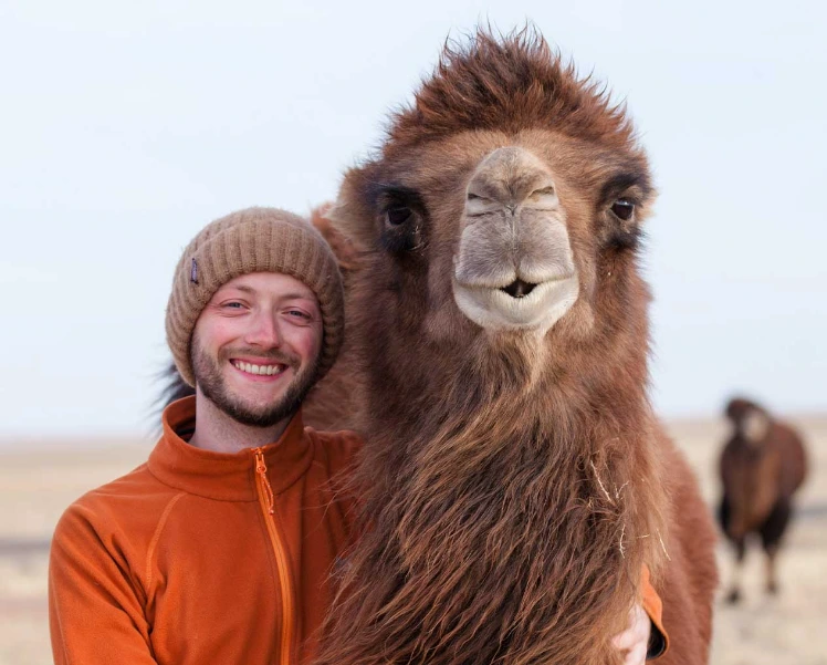 Malte 2019 in der Mongolei Junger Mann steht lachend neben einem Kamel